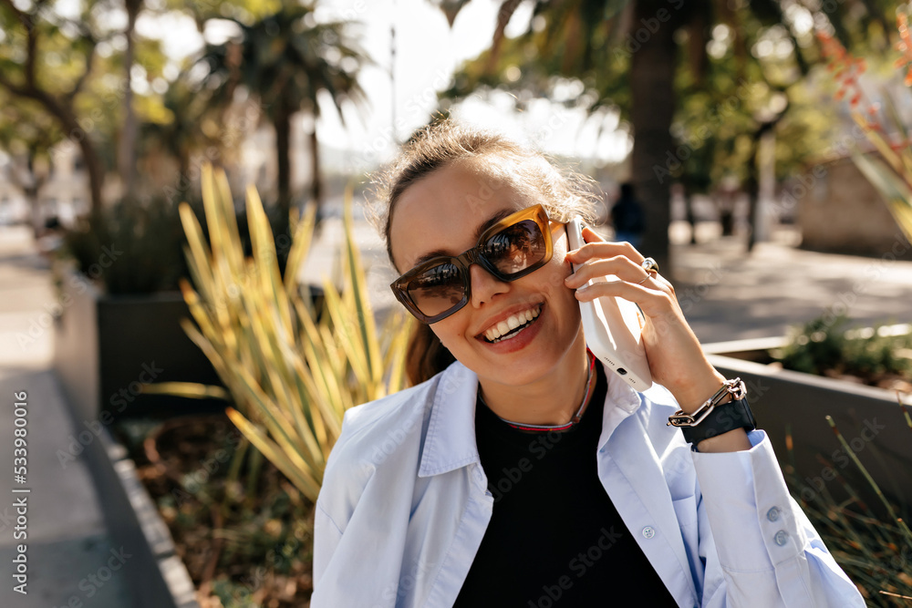 Attractive charming woman with gentle smile wearing black t-shirt and blue shirt in sunglasses sitting outdoor in sunlight and talking on the phone Attractive charming woman with gentle smile wearing black t-shirt and blue shirt in sunglasses sitting outdoor in sunlight and talking on the phone