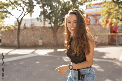 Excited lovely stylish woman with happy smile and loose wavy hair wearing black t-shirt and denim shorts walking around summer city in sunlight