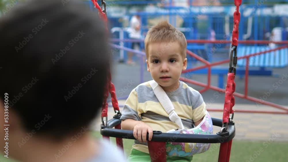Little blond 2 years old boy with a sad expression on his face, with broken arm in bandage outdoors sits alone on the swing at the playground. The worst summer leisure. Concept of health and medical. Little blond 2 years old boy with a sad expression on his face, with broken arm in bandage outdoors sits alone on the swing at the playground. The worst summer leisure. Concept of health and medical.