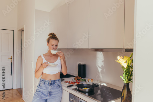 Young woman in top holding coffee cup near breakfast and lily flowers in kitchen.