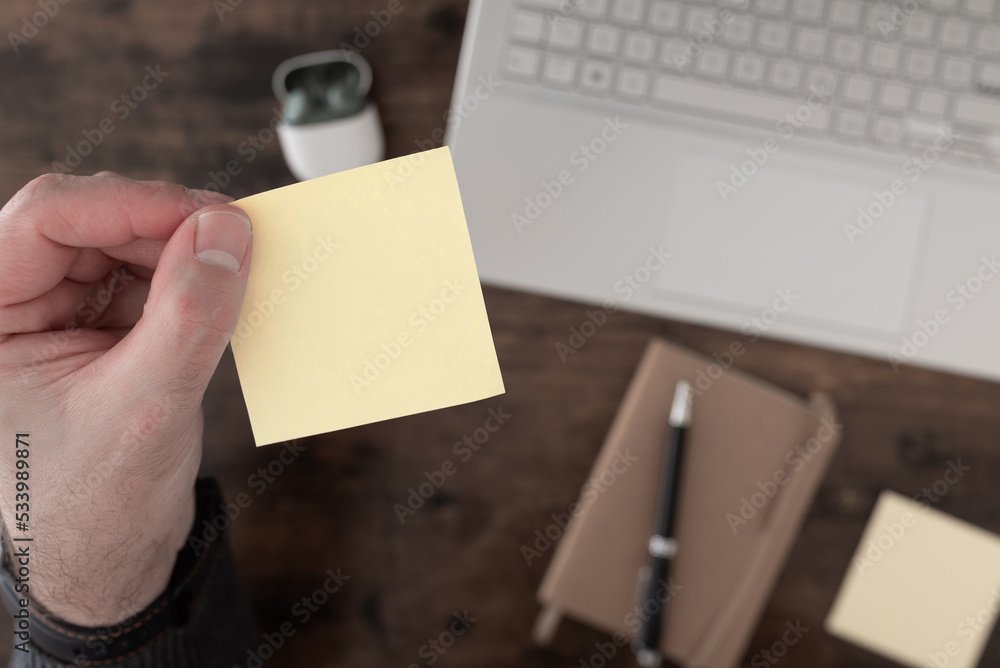 Obraz premium top down view of hand holding empty yellow sticky note above desk with laptop computer and notepad