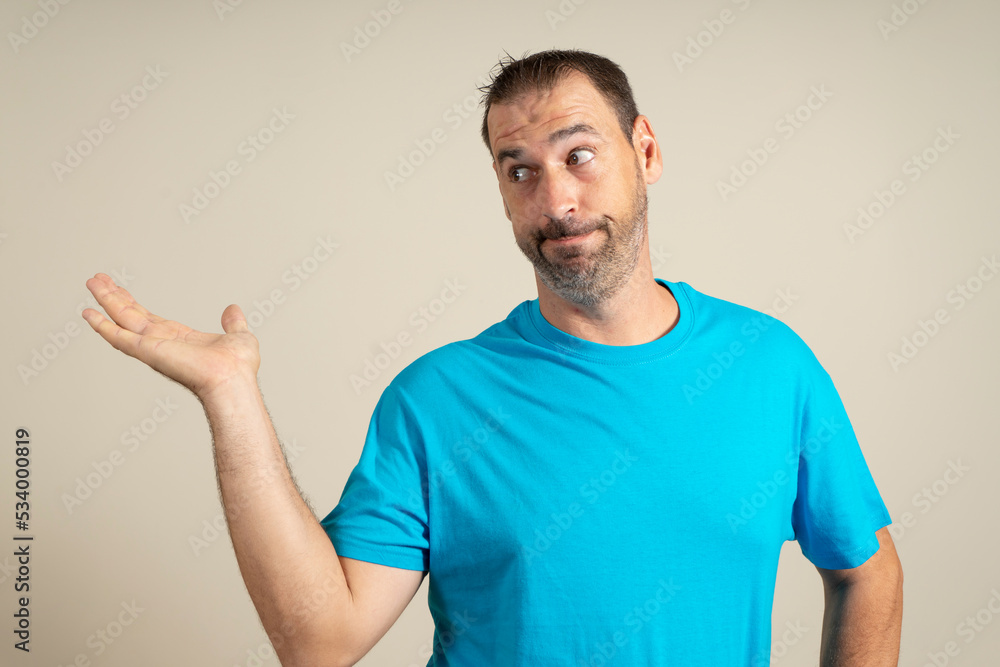 Hispanic man with a beard wearing a blue t-shirt in a skeptical attitude offering something in the palm of his hand, isolated over beige background. Hispanic man with a beard wearing a blue t-shirt in a skeptical attitude offering something in the palm of his hand, isolated over beige background.