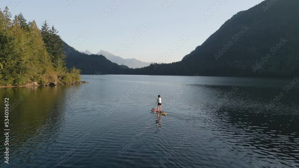 Adventurous Woman Paddling on a Paddle Board in a peaceful lake. Sunny Sunset. Hicks Lake, Sasquatch Provincial Park near Harrison Hot Springs, British Columbia, Canada. Slow Motion Adventurous Woman Paddling on a Paddle Board in a peaceful lake. Sunny Sunset. Hicks Lake, Sasquatch Provincial Park near Harrison Hot Springs, British Columbia, Canada. Slow Motion