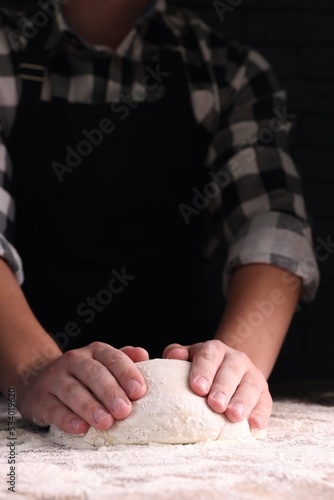 Man kneading dough at wooden table on dark background, closeup