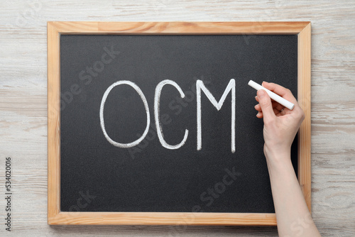 Woman writing abbreviation OCM (Organizational Change Management) on small blackboard with at white wooden table, top view