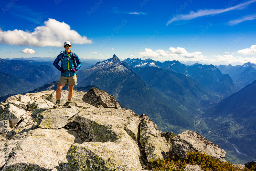 Adventurous athletic female hiker standing on top of a rugged mountain looking at the camera smiling with jagged mountains in the background in the Pacific Northwest.