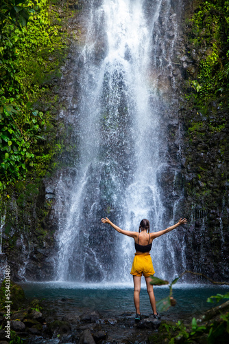 brave girl stands in front of a mighty waterfall with her hands raised in the air  celebration of a successful climb over a waterfall  tropical waterfall in Costa Rica  hidden gems of costa rica © Jakub