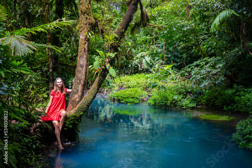 beautiful girl in red dress sitting on a tree by the tropical blue river in costa rica  volcano tenorio national park  blue river