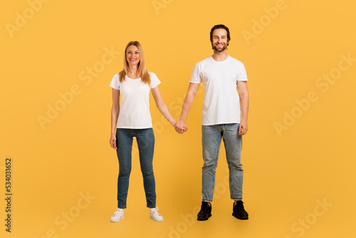 Smiling young caucasian female and male in white t-shirts hold hands, enjoy spare time, look at camera