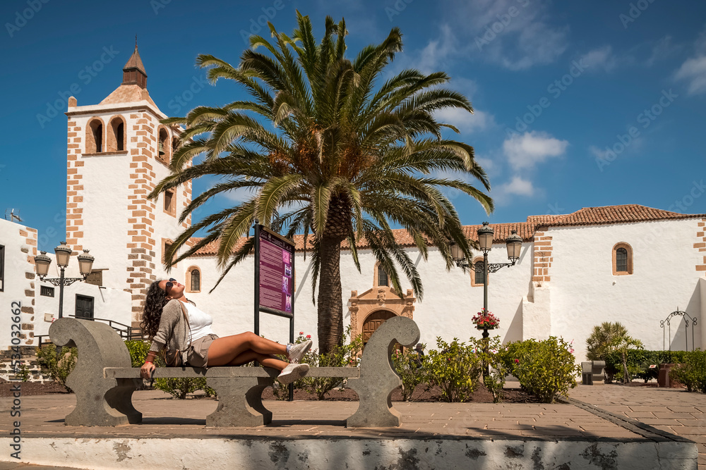 beautiful woman with curly hair lying on a bench in the main square of Betancuria, the ancient capital of fuerteventura, behind there is the ancient church with a white stone bell tower