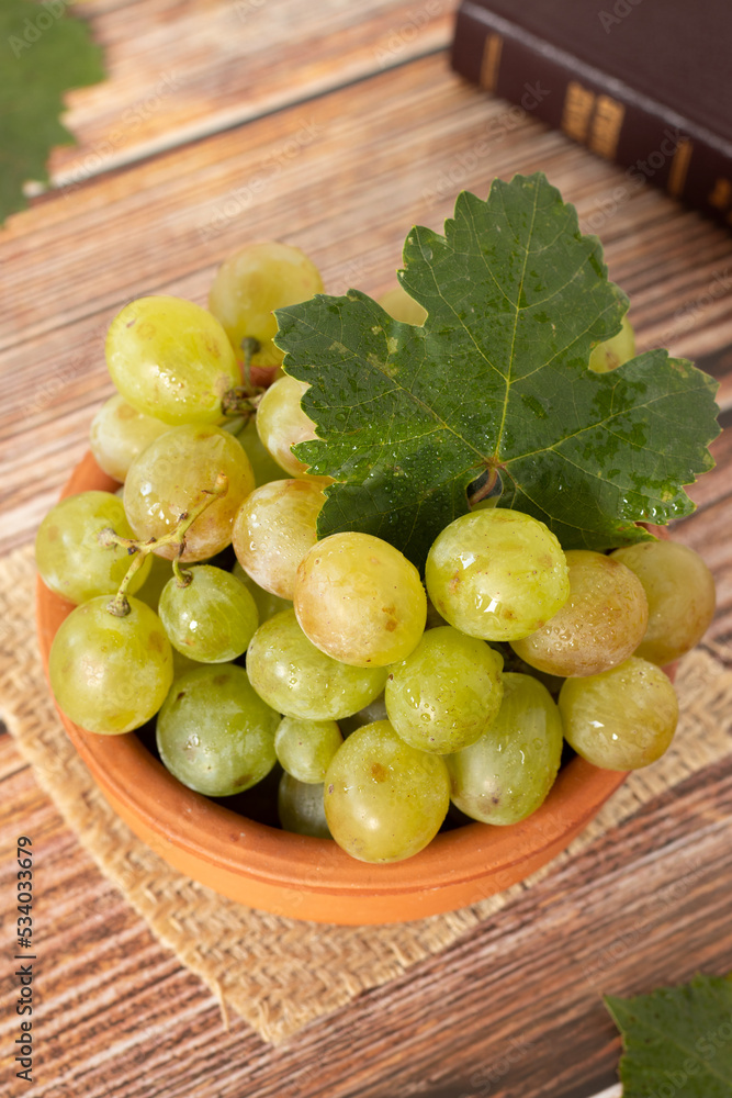 Bunch of green grapes with fresh vine leaf on a bowl with closed Holy Bible on wooden table. Vertical shot. A closeup. Bunch of green grapes with fresh vine leaf on a bowl with closed Holy Bible on wooden table. Vertical shot. A closeup.