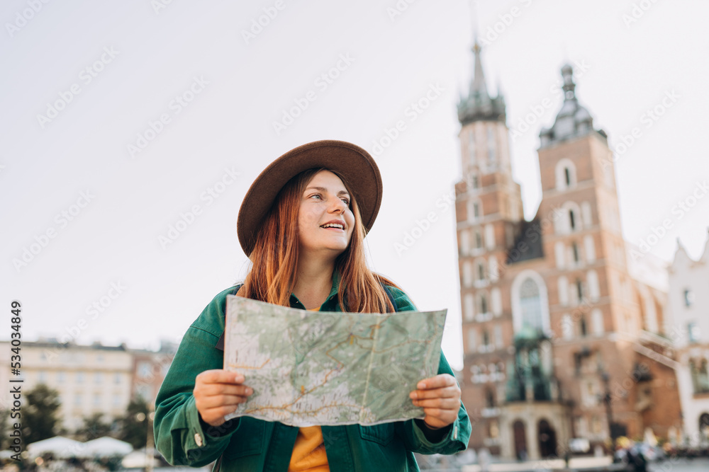 Attractive young female tourist is exploring new city. Redhead girl holding a paper map on Market Square in Krakow. Traveling Europe in autumn. St. Marys Basilica