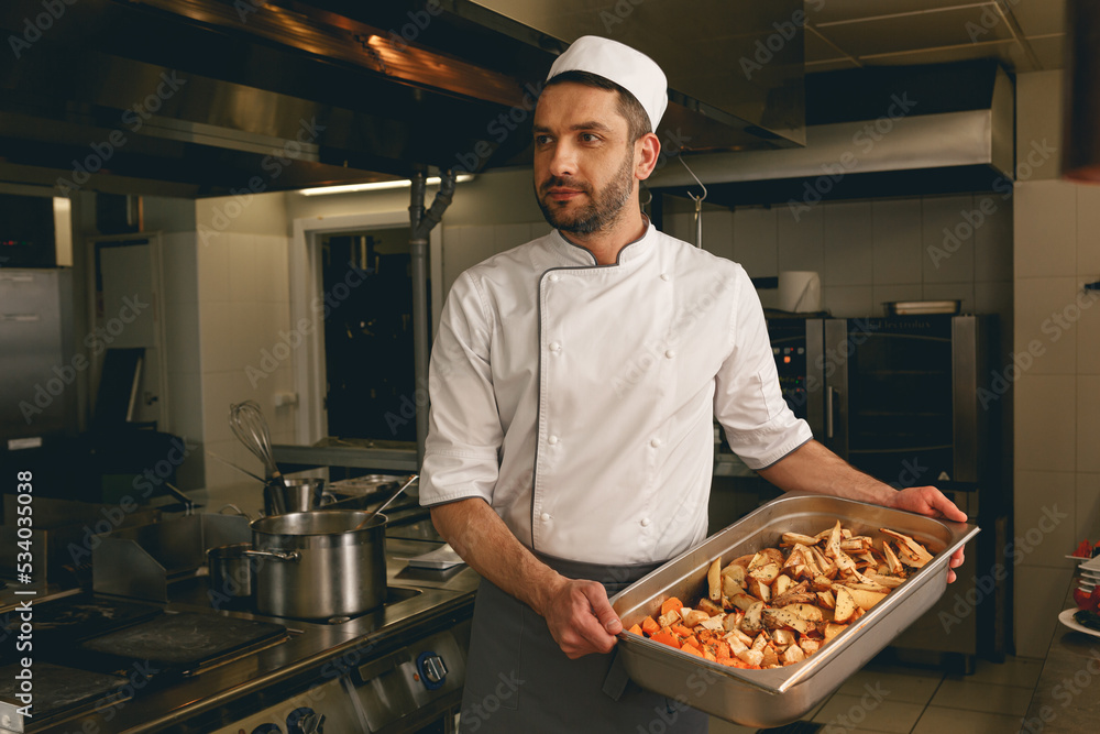 Chef holding tray with baked vegetables while standing in the kitchen of restaurant