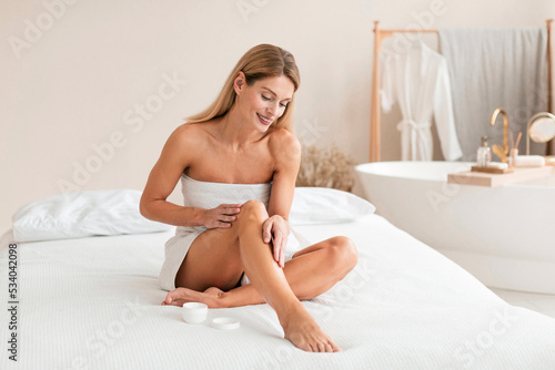 Happy middle aged lady applying moisturizer cream on legs, caring for body and moisturizing skin after shaving photo