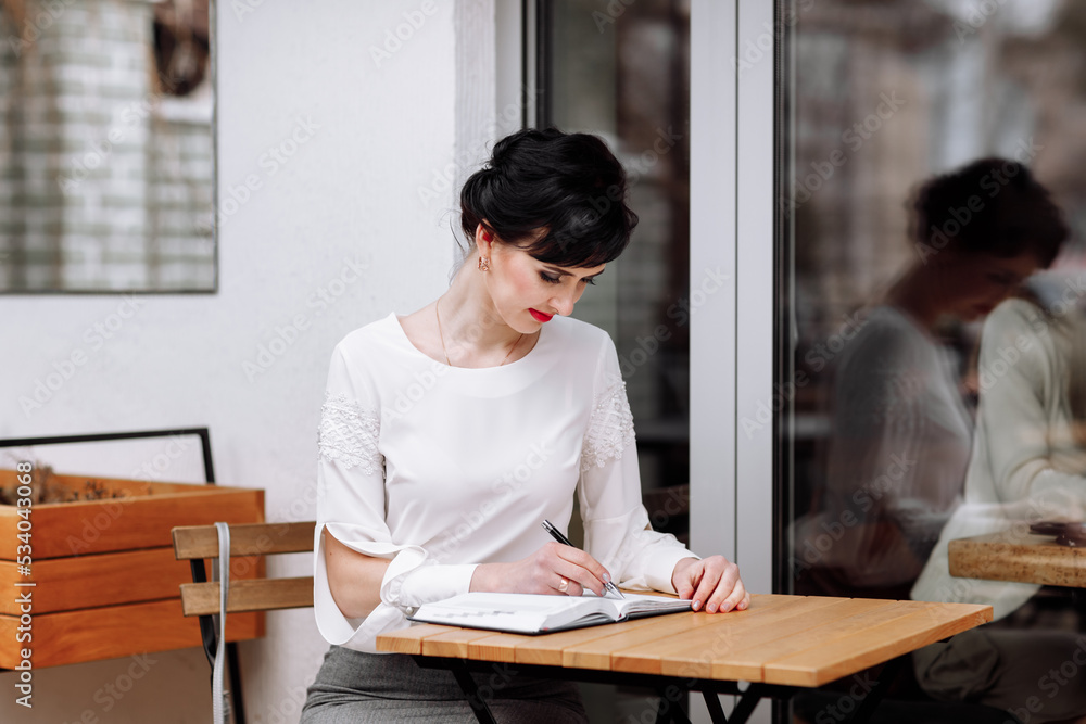 Serious focused smart businesswoman is writing notes on notepad, making agenda on personal organizer. Female in business suit sitting at table at cafe on terrace, work and event planning. Serious focused smart businesswoman is writing notes on notepad, making agenda on personal organizer. Female in business suit sitting at table at cafe on terrace, work and event planning.