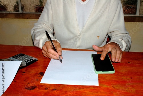 El profesional médico escribe una receta en su oficina en la mesa de madera lustrada haciendo cálculos con la tecnología mòvil,  forma un diseño de estilo de vida con un fondo de la ventana 