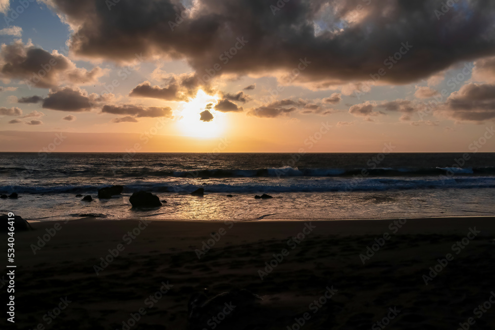 Fototapeta premium Scenic view during sunset on the volcanic sand beach Playa del Ingles in Valle Gran Rey, La Gomera, Canary Islands, Spain, Europe. Sun beam reflection on water surface. Calm atmosphere at the seaside