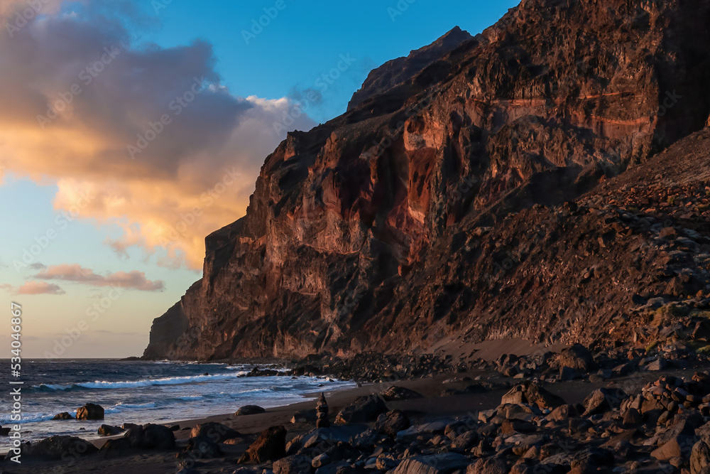 Scenic view during sunset on the volcanic sand beach Playa del Ingles in Valle Gran Rey, La Gomera, Canary Islands, Spain, Europe. Massive cliffs of the La Mercia range. Calm atmosphere at the seaside