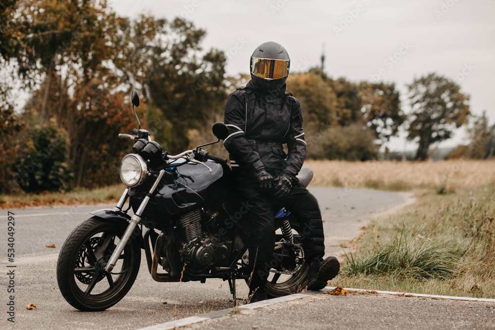 male motorcyclist in protective uniform and helmet with custom motorcycle cafe racer in autumn on the road.