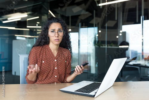 Frustrated and sad business woman with phone in hands looking at camera, latin american woman working with laptop inside modern office building, unclear emotional state of female employee.