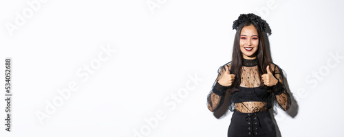 Image of happy asian woman in halloween party dress showing thumbs-up in approval. Girl wearing as witch or widow looking pleased, like or agree with something, white background