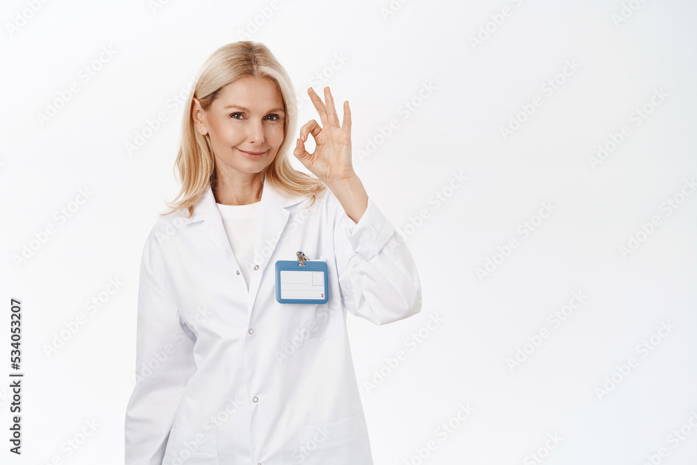 Excellent. Smiling beautiful woman doctor, senior lady in medical uniform, shows okay, ok sign, approves smth, agrees, stands over white background Excellent. Smiling beautiful woman doctor, senior lady in medical uniform, shows okay, ok sign, approves smth, agrees, stands over white background