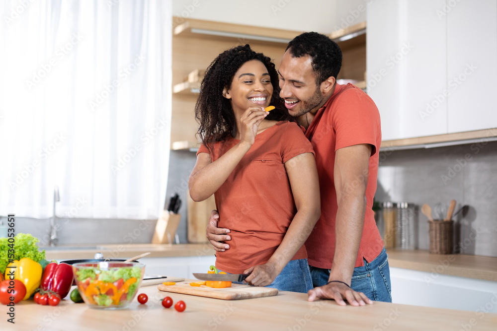 Cheerful millennial black husband hugging lady, woman cutting organic vegetables at salad and feeding man