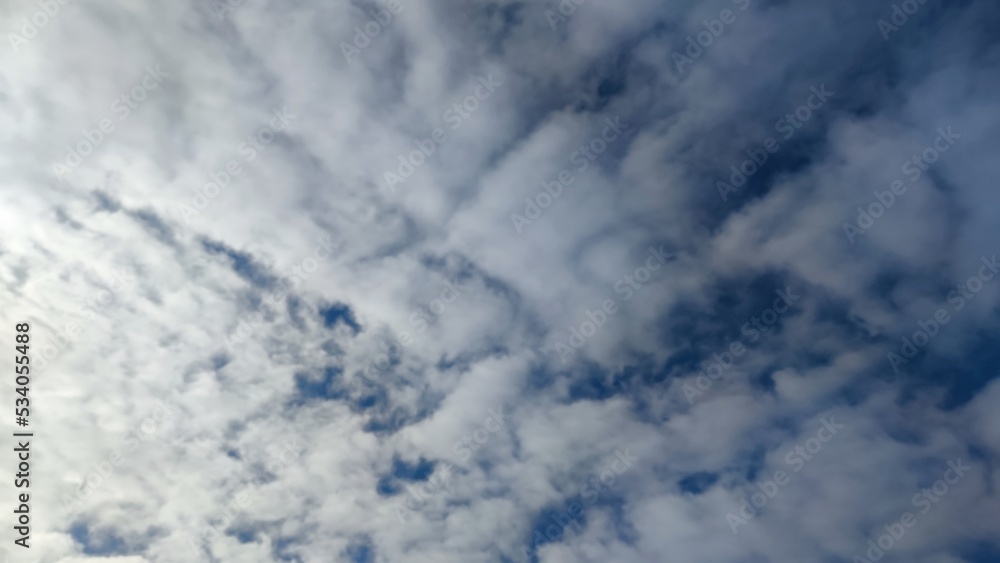 The sun shines behind the clouds. Clear day in the sky high cumulus clouds. They are white in different shapes and sizes. Behind the clouds, the sun shines brightly, shining through the clouds. The sun shines behind the clouds. Clear day in the sky high cumulus clouds. They are white in different shapes and sizes. Behind the clouds, the sun shines brightly, shining through the clouds.