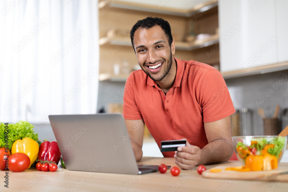 Cheerful millennial black male at table with organic vegetables uses laptop and credit card Cheerful millennial black male at table with organic vegetables uses laptop and credit card