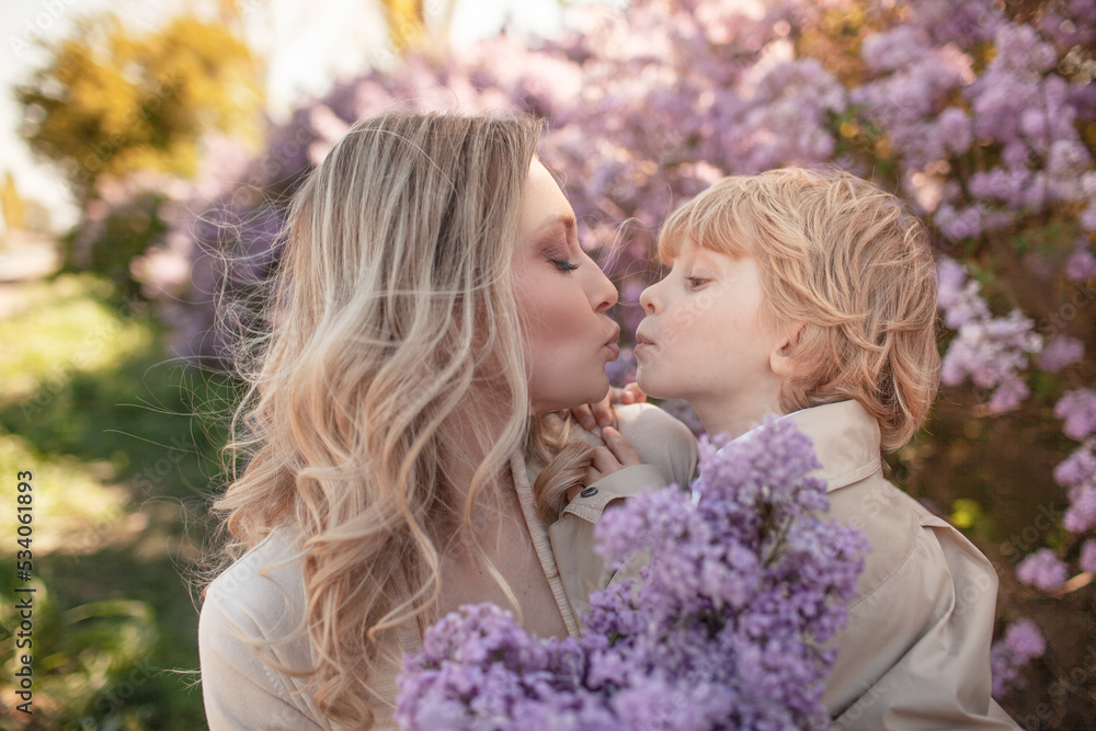 A little stylish boy with mother walks among flower trees in spring and mom holds a kid and hugging outdoor in the nature A little stylish boy with mother walks among flower trees in spring and mom holds a kid and hugging outdoor in the nature