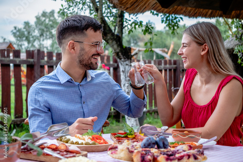 Happy young romantic couple toasting with shots of brandy with a particular narrow neck glasses in a beautiful traditional outdoor country restaurant.
