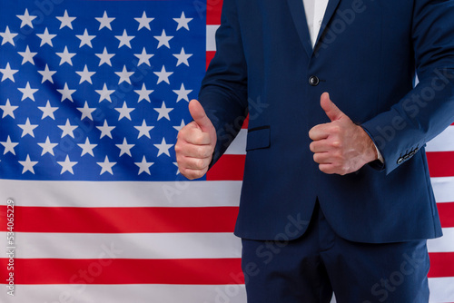 A businessman with thumbs up in victory sign with the United States flag in the background. Victory for the USA. Support for U.S. policy