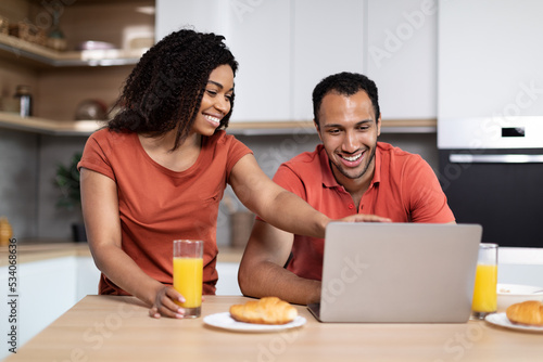 Glad millennial black family enjoy breakfast in morning together, lady pointing finger at laptop