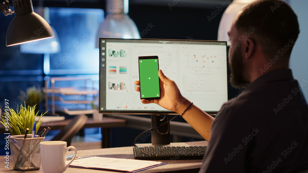Project manager looking at greenscreen template on smartphone display, working at night. Holding mobile phone with isolated mockup background, blank chromakey and copyspace. Tripod shot.