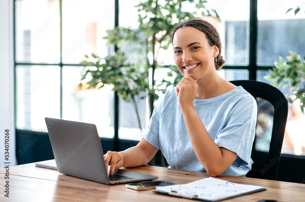 Confident successful elegant pretty brazilian or hispanic young woman, company ceo, product or financial manager, in blue shirt, sits at work desk in modern office, looking at camera, smiles friendly