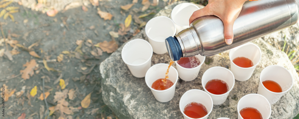 Close-up top above view woman hand hold steel thermos pouring hot tea many  white paper cup group picnic mountain trekking outdoors fall sunny morning  evening. Person thermo flask warm drink sharing Stock