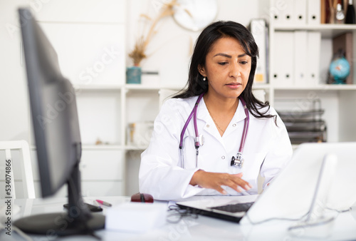 Latin american woman experienced physician filling up medical forms on laptop while sitting at table in office.