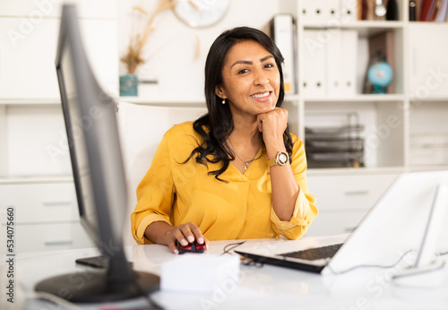 Portrait of confident smiling latin american female office employee during daily work with computer photo