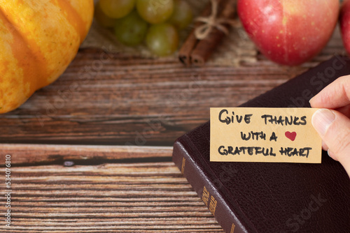 Hand holding a handwritten thanksgiving note to give thanks with a grateful heart with closed Holy Bible Book and fresh autumn fruit on wooden background with copy space. A closeup. Christian concept.