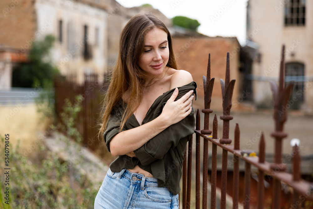 Gorgeous female model with long hair, dressed in jeans and shirt, posing against wrought-iron fence and building on background