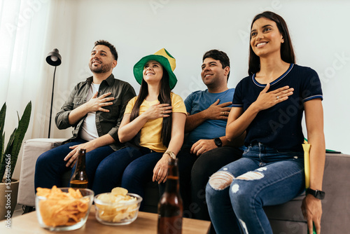 Multiethnic group of friends listening and singing Brazilian national anthem before watching sports championship on TV together at home