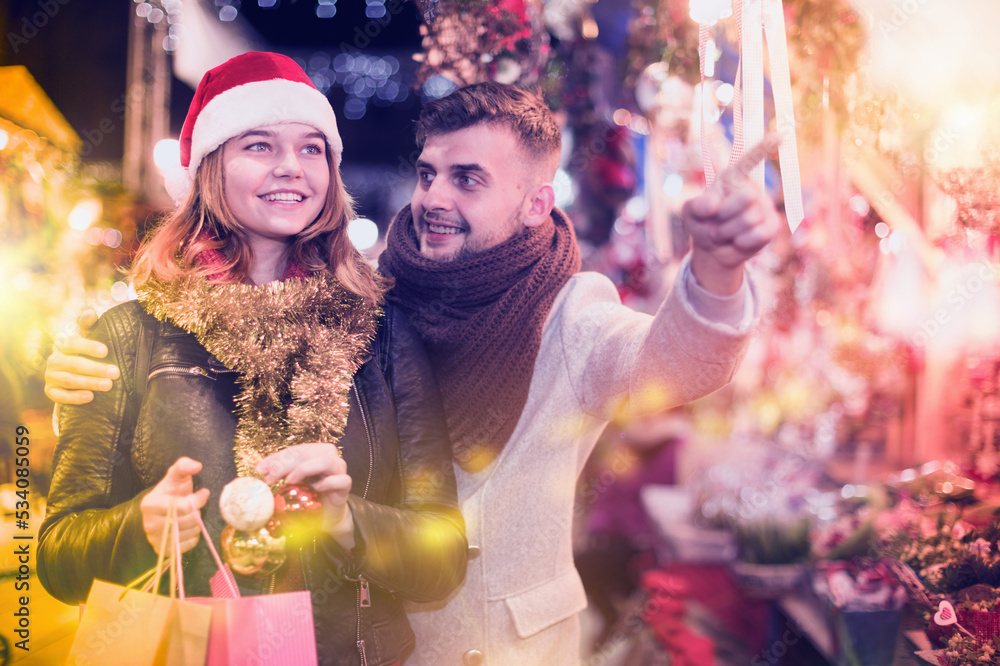 Joyful young couple in hat buying decoration at Christmas Fair, man points to decorations