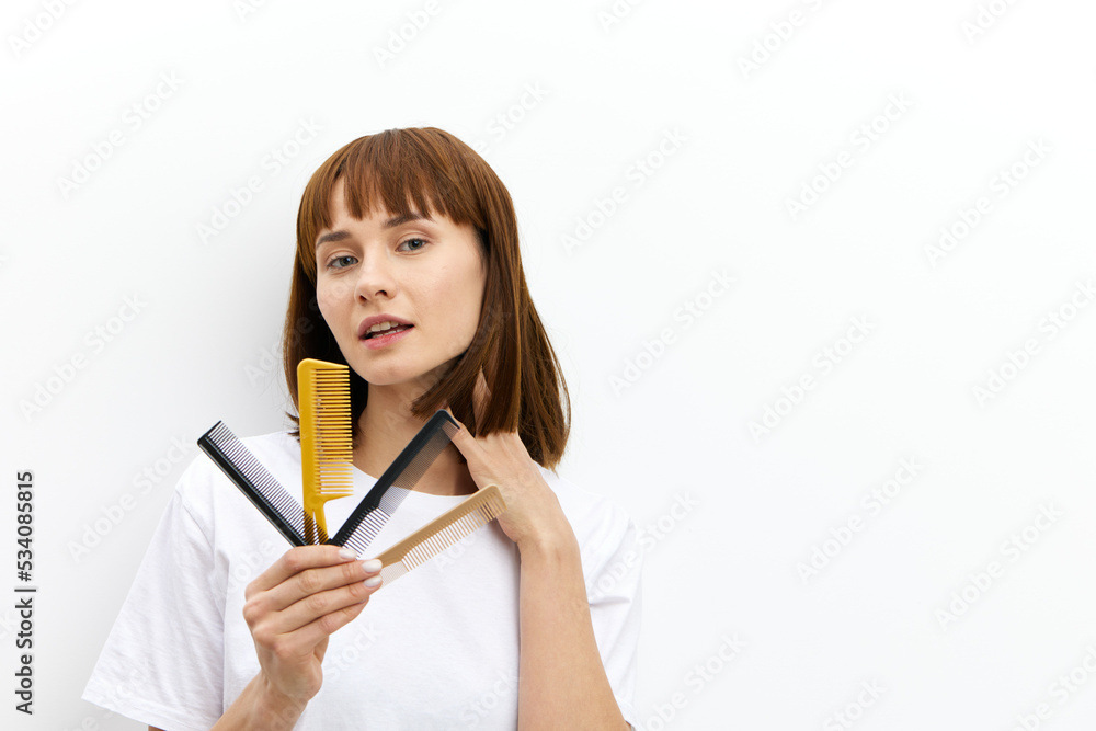 a sweet, pleasant woman with a beautiful hairstyle holds a lot of different combs in one hand, and touches her head with the other, standing in a white T-shirt on a white background a sweet, pleasant woman with a beautiful hairstyle holds a lot of different combs in one hand, and touches her head with the other, standing in a white T-shirt on a white background