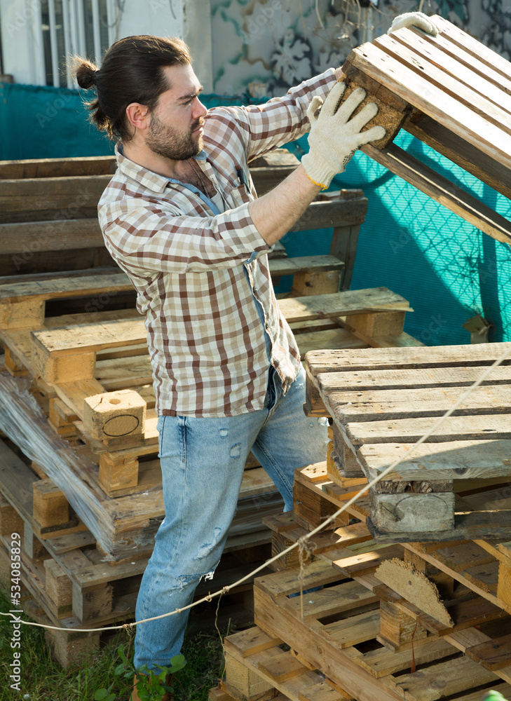 Young man in work wear moving wooden pallets at garden