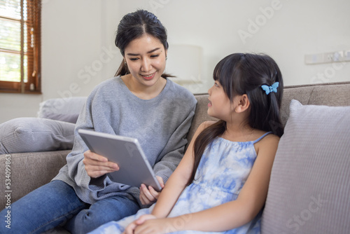 Happy asian mom holding her daughter playing and reading E-book on sofa at home.little girl hugging her mother smile and love having fun at home.Mother day concept