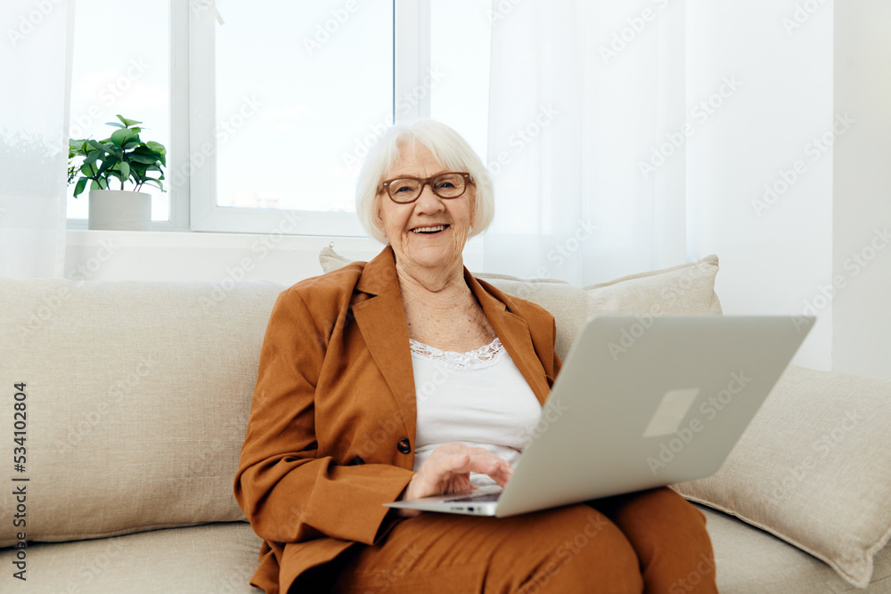 a happy elderly woman is sitting looking at a laptop on the sofa in a relaxed pose in a bright room near the window with glasses on her face and working remotely smiling broadly with pleasure a happy elderly woman is sitting looking at a laptop on the sofa in a relaxed pose in a bright room near the window with glasses on her face and working remotely smiling broadly with pleasure