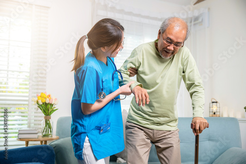 International Day for the Elderly. Young carer supporting helping senior man stand up with walking stick from sofa, Nurse wearing blue uniform help helping his patient senior old man with walking cane