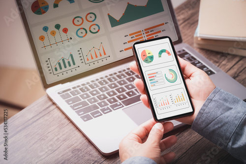 Businesswoman looking graph dashboard analysis performance on smartphone and laptop computer ,using mobile phone for update performance data dashboard graph with dark background.