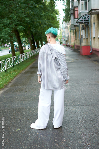 A woman with green hair in white clothes and with an umbrella walks down the street after the rain
