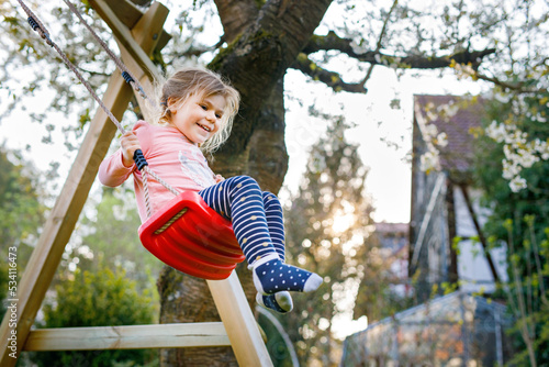 Happy beautiful little toddler girl having fun on swing in domestic garden. Cute healthy child swinging under blooming trees on sunny spring day. Baby laughing and crying